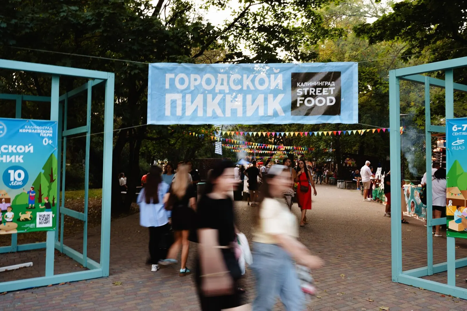 Вход на площадку фестиваля «Городской пикник» в парке: баннер над проходом, гирлянды из флажков, прохожие